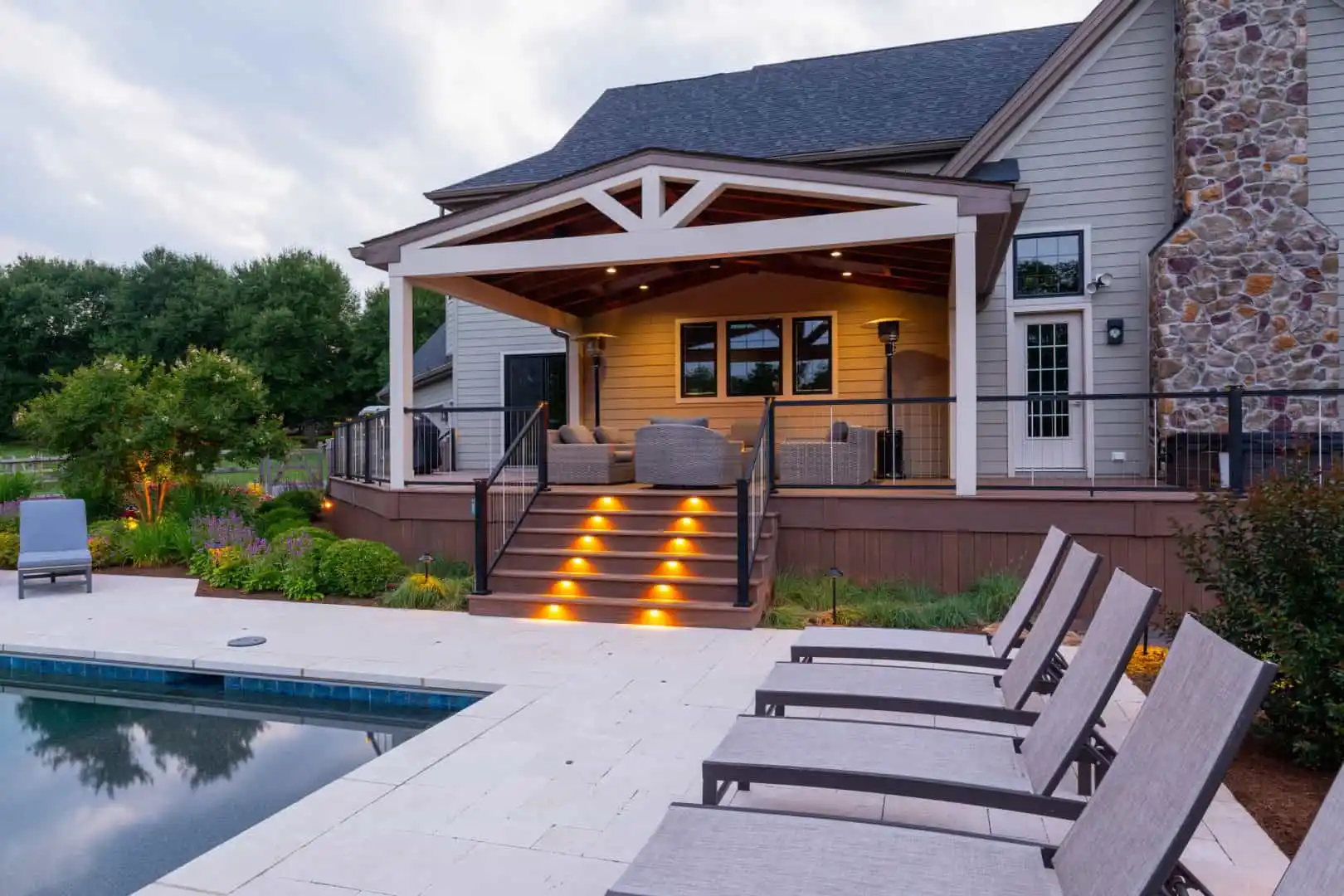 A modern house with a covered patio, illuminated stairs, and outdoor seating overlooks a pool lined with lounge chairs, surrounded by greenery and landscaping at dusk.