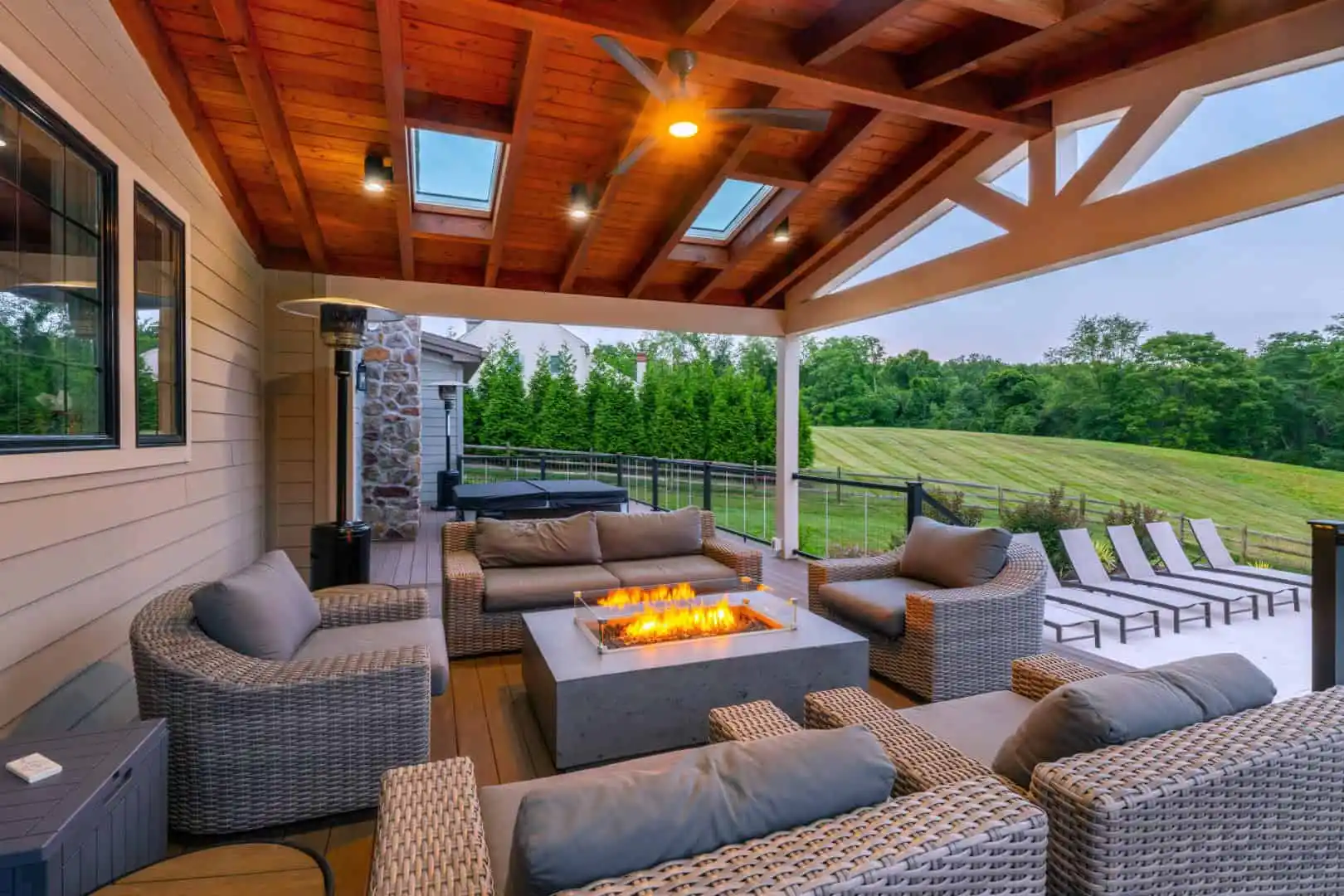 A cozy outdoor patio features wicker furniture with gray cushions, centered around a modern fire pit. A wooden pergola roof with skylights covers the area. This serene outdoor living setup in Delaware County, PA, is complemented by a manicured lawn and trees, with lounge chairs on a nearby deck.