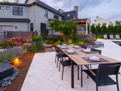 A modern backyard epitomizes outdoor living in Delaware County, PA, with a wooden dining table set on a tiled patio. Black chairs encircle it, while a landscaped garden bursting with flowers complements the pool and lounge chairs. A spacious house looms in the background under a cloudy sky.