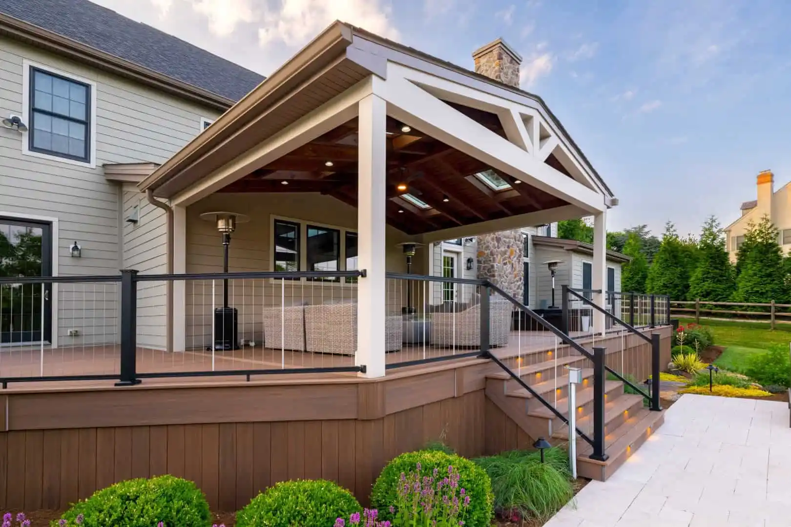 A spacious, elevated wooden deck with a covered seating area is attached to a beige house in Delaware County, PA. The deck features a glass railing, stairs, and a stone fireplace. Surrounding the deck are neatly landscaped shrubs and flowers, showcasing expert landscaping services.