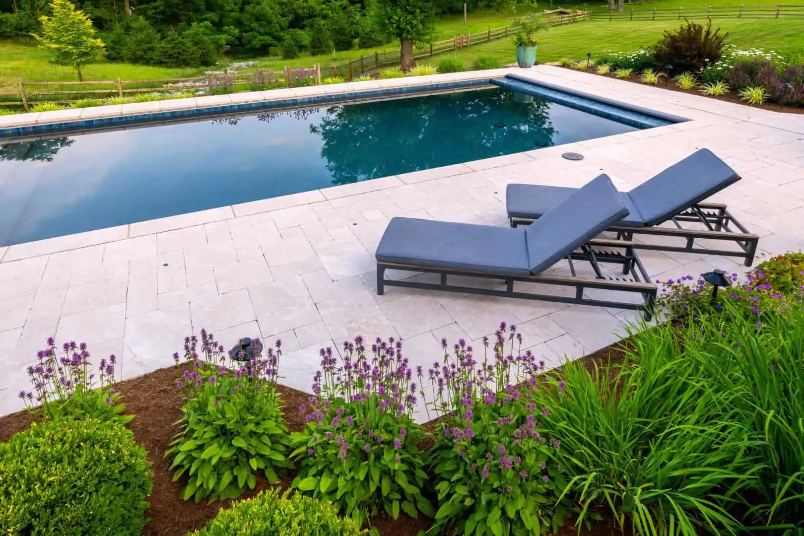 A serene backyard pool area with two gray lounge chairs on a stone patio showcases the beauty of outdoor living in Delaware County, PA. Purple flowers and lush green plants border the pool, framed by trees and a wooden fence, illustrating expert landscaping services.