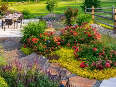 A landscaped garden with vibrant red and pink flowers is surrounded by lush greenery in Delaware County, PA. A wooden fence encloses the area, where a patio with a table and chairs offers the perfect outdoor living experience, set against a sunny, green lawn backdrop.