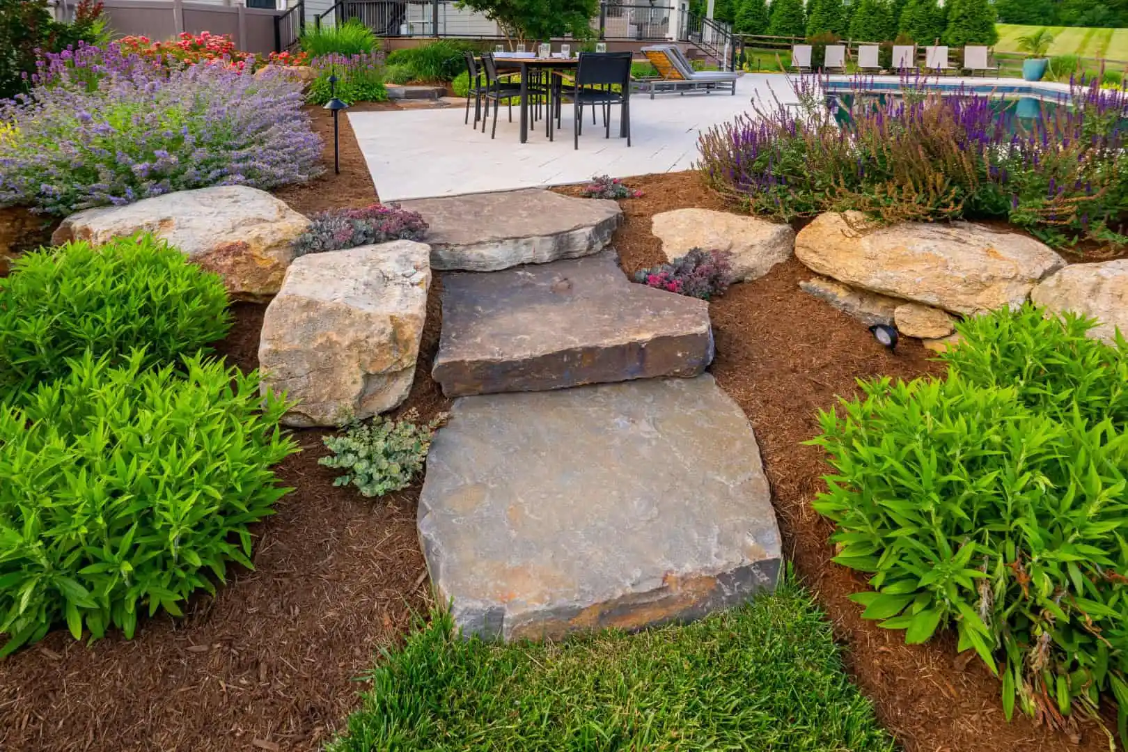 Three large stone steps lead to a charming patio area with chairs and tables. The path is bordered by lush greenery and vibrant flowers, including purple and pink blooms. Mulch covers the surrounding garden areas, enhancing the colorful landscape, thanks to expert landscaping services in Delaware County, PA.