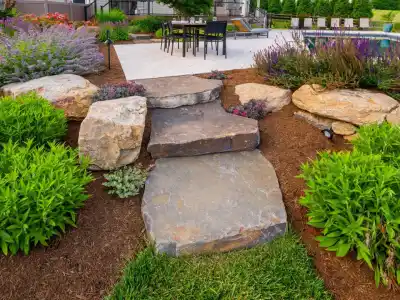 Three large stone steps lead to a charming patio area with chairs and tables. The path is bordered by lush greenery and vibrant flowers, including purple and pink blooms. Mulch covers the surrounding garden areas, enhancing the colorful landscape, thanks to expert landscaping services in Delaware County, PA.