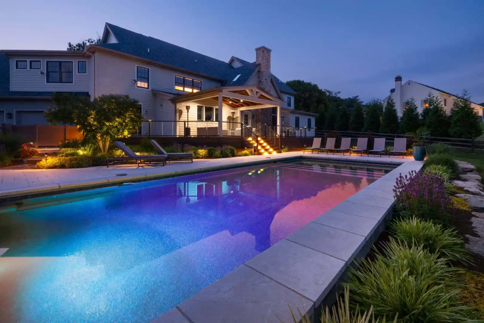 Evening view of a lit outdoor swimming pool casting a multicolored glow, perfect for outdoor living in Delaware County, PA. Deck chairs and landscaped greenery enhance the ambiance. In the background, a large house with illuminated windows and a covered porch completes this enchanting scene.