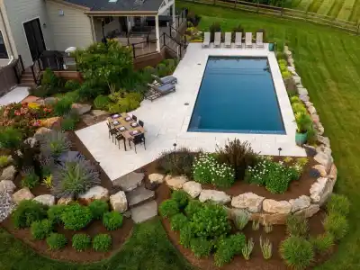 Aerial view of a backyard with a rectangular pool, surrounded by an expertly crafted stone patio, showcases premium hardscaping services in Delaware County, PA. Lounge chairs and a dining set complement the lush gardens adorned with colorful plants and rocks encircling the area.