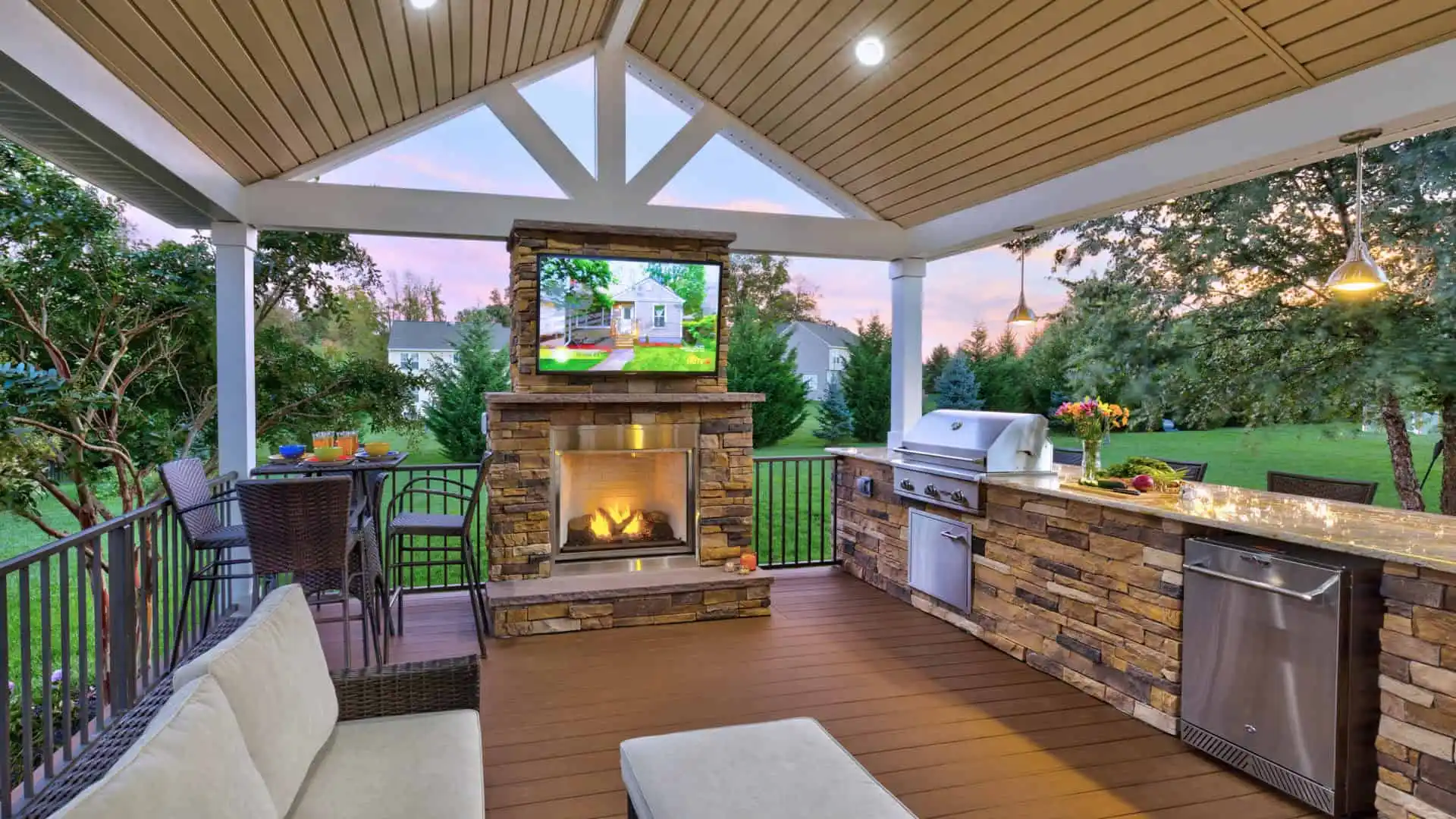 The outdoor patio, a gem of outdoor living in Delaware County, PA, showcases a covered seating area with a sofa, a stone fireplace topped with a TV, and amenities like a built-in grill and fridge. The bar with stools provides the perfect spot to admire the lush garden at sunset.