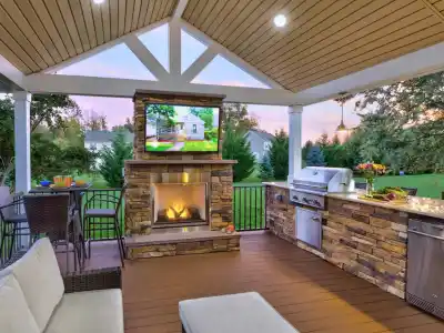 The outdoor patio, a gem of outdoor living in Delaware County, PA, showcases a covered seating area with a sofa, a stone fireplace topped with a TV, and amenities like a built-in grill and fridge. The bar with stools provides the perfect spot to admire the lush garden at sunset.
