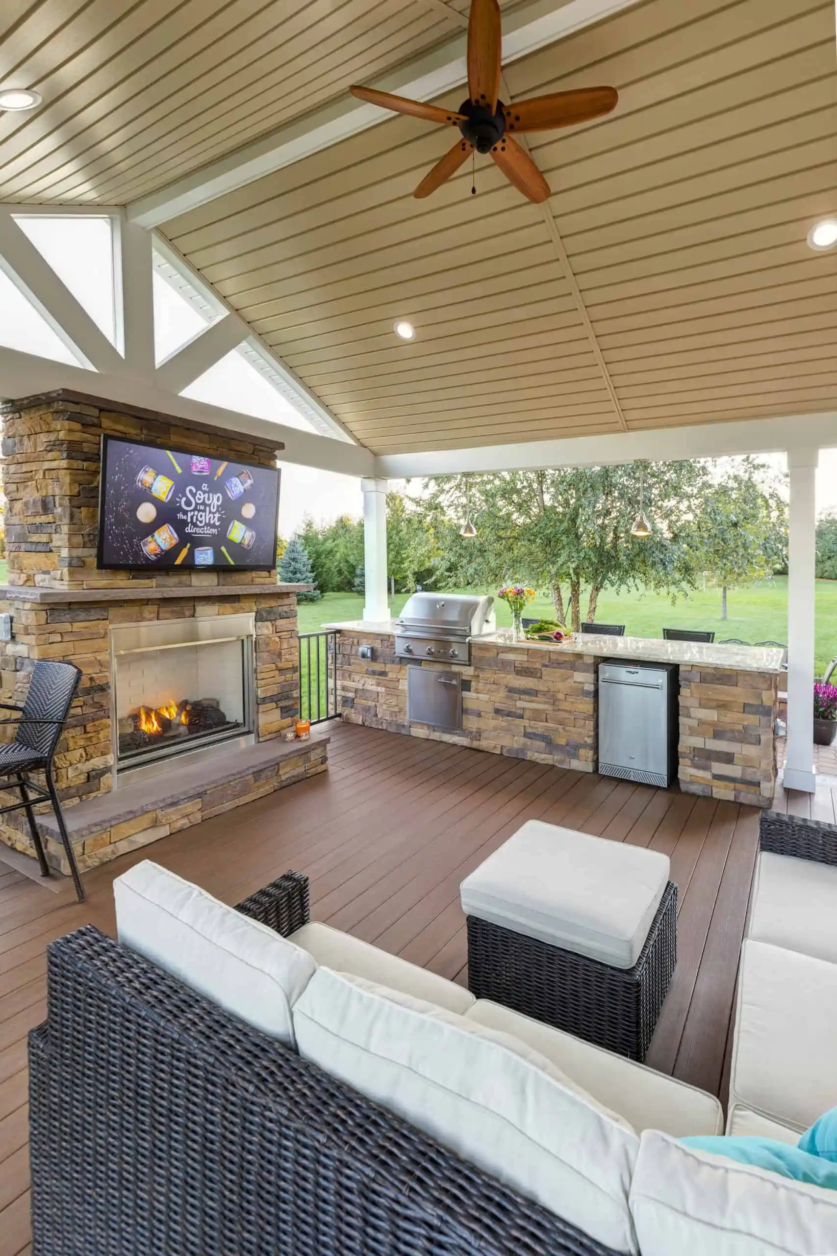 A covered outdoor patio features a wicker sofa, fireplace, TV, built-in grill, mini fridge, and sink. Ceiling fan and recessed lights hang above. The patio overlooks a green lawn with trees in the background.