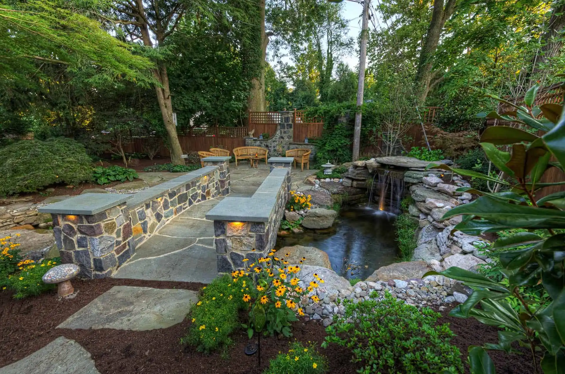 A landscaped backyard features a stone patio with seating, surrounded by lush greenery, yellow flowers, a small pond, and a waterfall, creating a peaceful outdoor retreat.