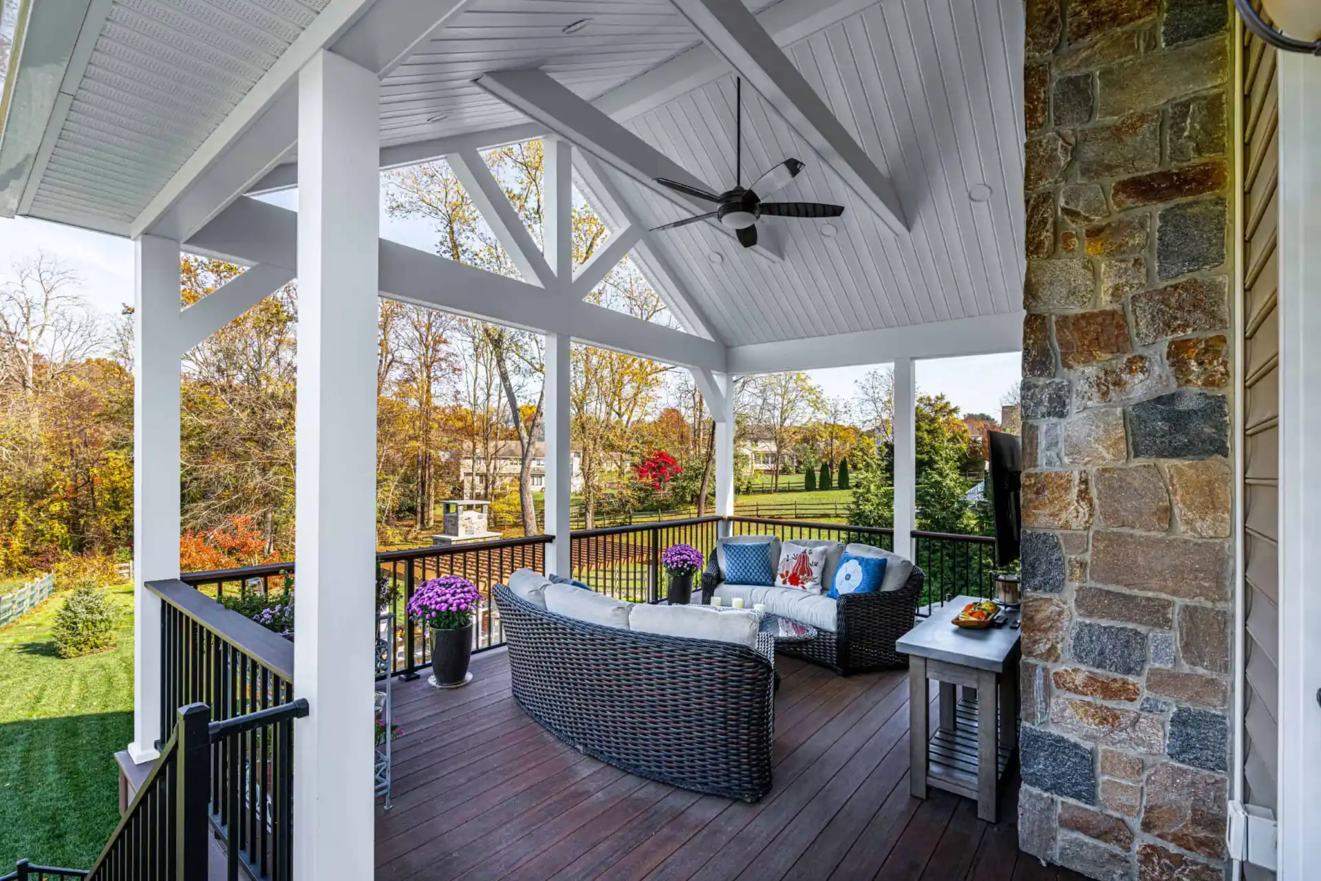 Covered patio with wicker furniture and colorful cushions overlooking a lush lawn and trees, perfect for enhancing your outdoor living in Delaware County, PA. Ceiling fans hang from a white roof with exposed beams, while a stone wall on the right and potted flowers complete this serene setting.