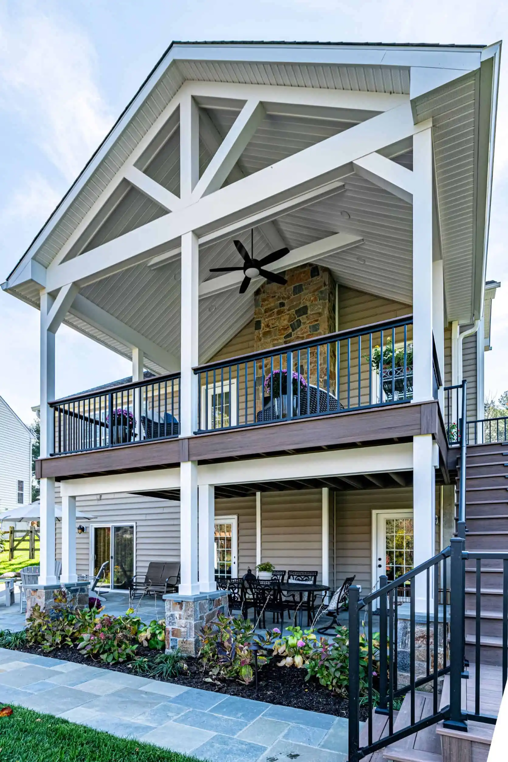 Elevated two-story porch in Delaware County, PA, with a gabled roof and white trusses. Upper level boasts outdoor seating and ceiling fan, while the lower level showcases a patio dining set. Stone and clapboard finish blend seamlessly with expert landscaping services and stairs leading up.