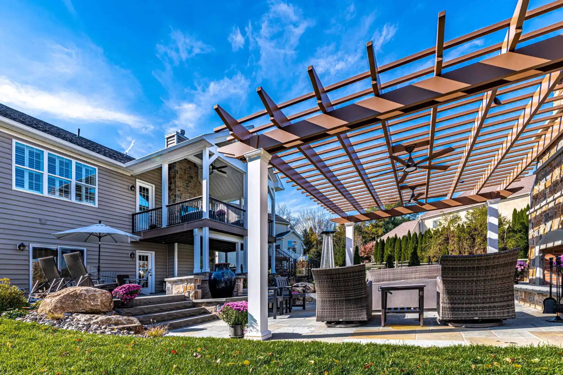 A modern backyard in Delaware County, PA, boasts a large house with a balcony and stone fireplace. The pergola-covered patio, adorned with outdoor furniture and vibrant flowers, showcases exquisite landscaping services. The sky is clear with a few clouds.