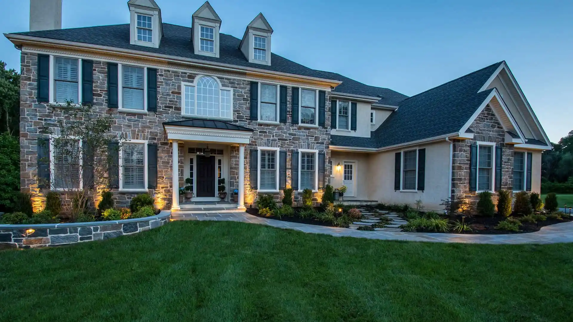 This large two-story stone and stucco home boasts a symmetrical facade with three dormer windows highlighted by exterior lighting. A white-columned portico marks the entrance, while expert hardscaping services in Delaware County, PA, have crafted the stone pathway through the neatly manicured lawn.