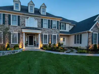 This large two-story stone and stucco home boasts a symmetrical facade with three dormer windows highlighted by exterior lighting. A white-columned portico marks the entrance, while expert hardscaping services in Delaware County, PA, have crafted the stone pathway through the neatly manicured lawn.