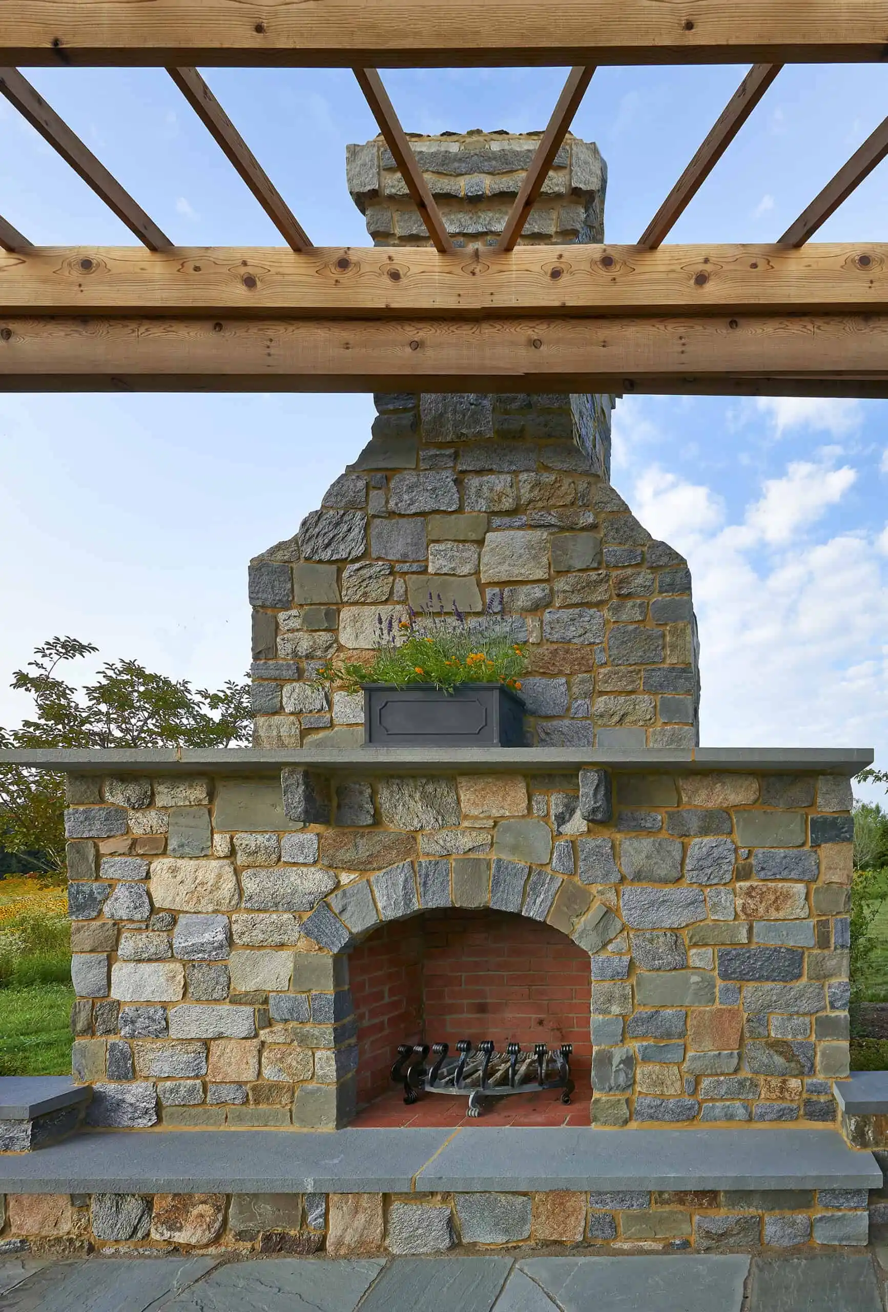A stone outdoor fireplace with a brick firebox sits beneath a wooden pergola. A rectangular planter with greenery is on the mantel, and the sky with clouds is visible in the background.