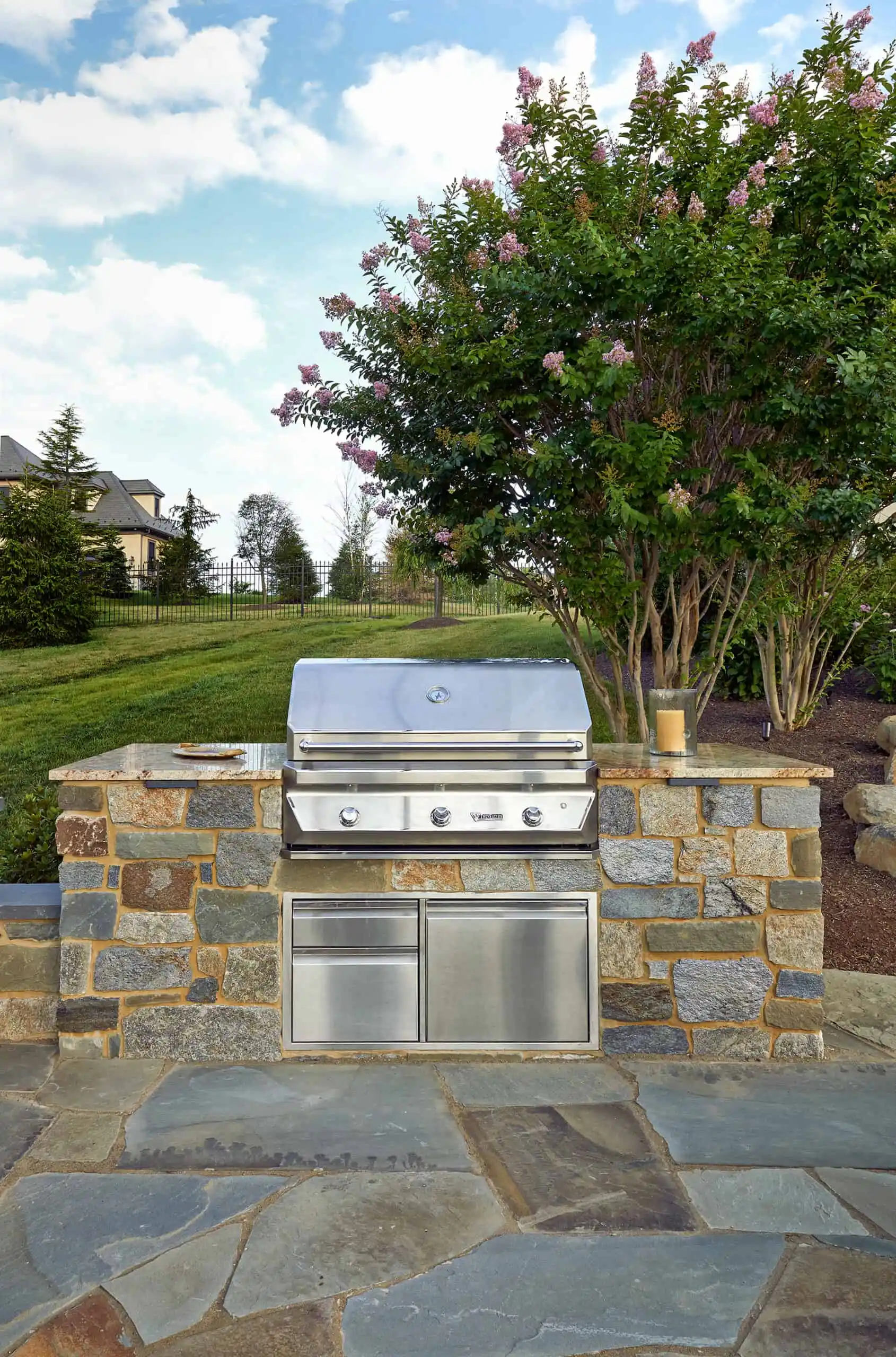 A built-in stainless steel grill set in a stone countertop outdoors, with a cup placed on top. Behind, there’s a tree with pink flowers and a well-kept lawn under a partly cloudy sky.