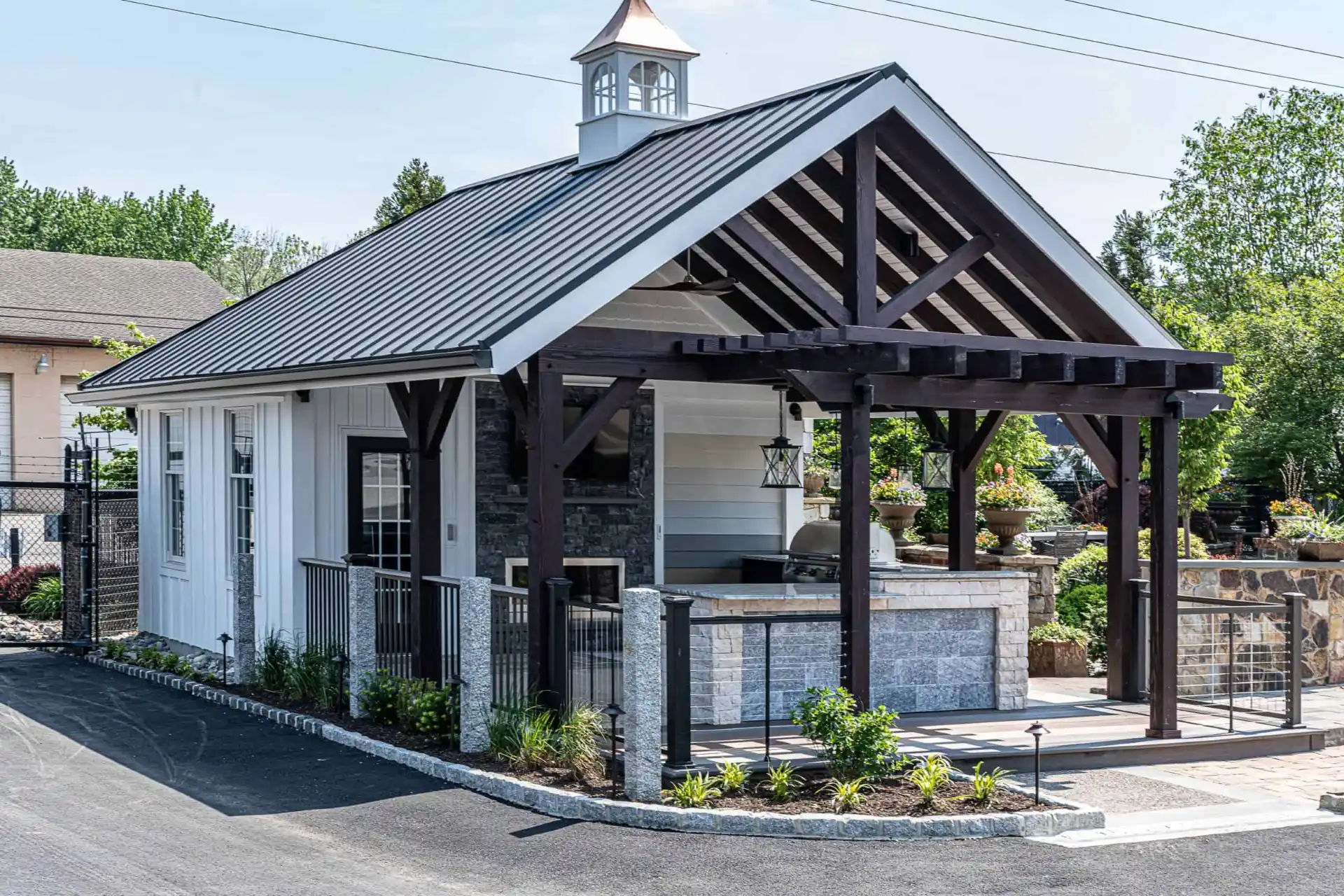 A small modern building with a black metal roof and wooden beams, featuring a cupola on top, is nestled in a beautifully landscaped area. It boasts a stone patio and decorative plants, exemplifying expert hardscaping services in Delaware County, PA. A paved driveway completes the inviting setting.