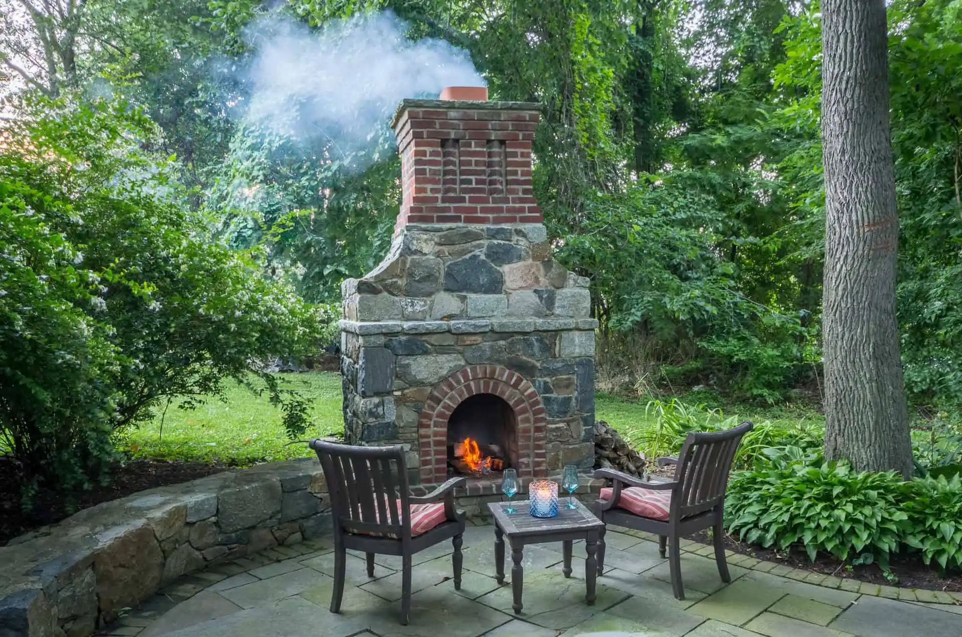 Outdoor stone and brick fireplace with a burning fire, two wooden chairs with cushions, and a small table on a stone patio surrounded by lush green trees and plants. Smoke rises from the chimney.