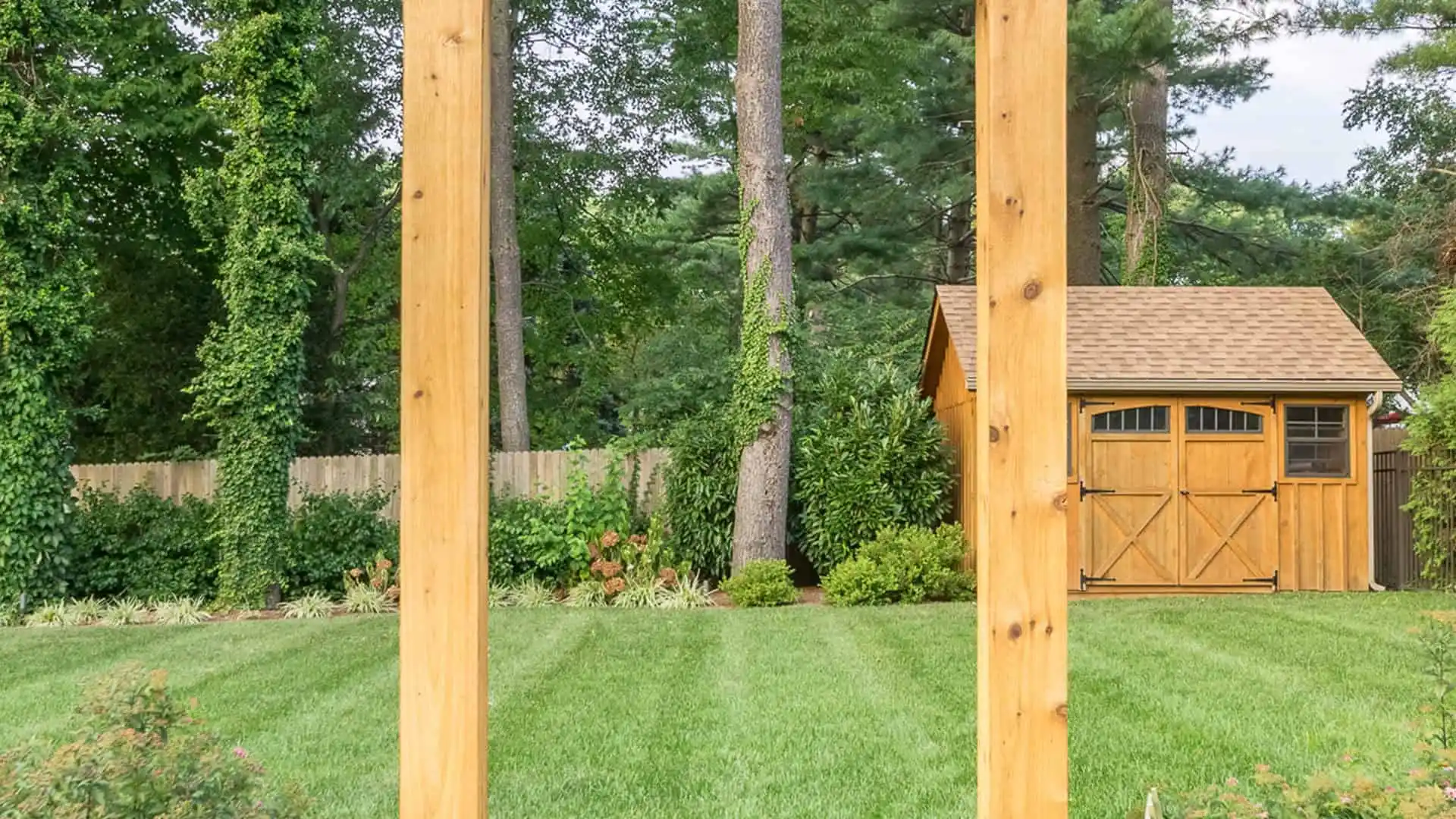 View of a backyard with a neatly mowed lawn and a wooden shed with double doors, framed by tall trees in the background. The foreground features two vertical wooden beams, highlighting the potential for exquisite outdoor living in Delaware County, PA.