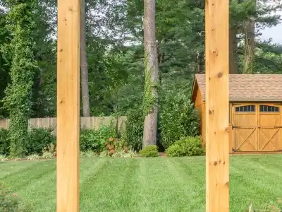 View of a backyard with a neatly mowed lawn and a wooden shed with double doors, framed by tall trees in the background. The foreground features two vertical wooden beams, highlighting the potential for exquisite outdoor living in Delaware County, PA.