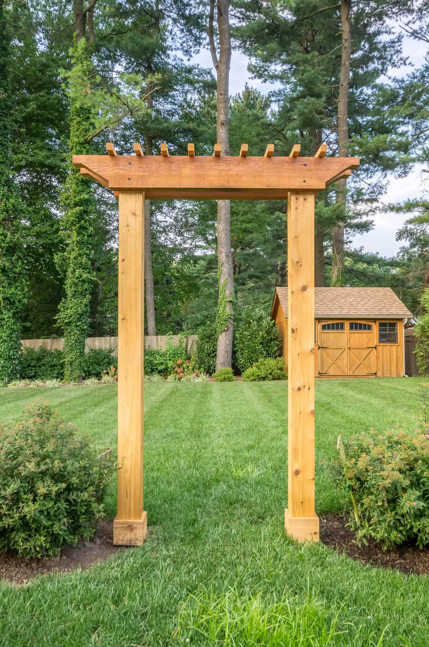 A wooden garden arbor stands on a grassy lawn with shrubs on either side; large trees and a wooden shed are visible in the background.