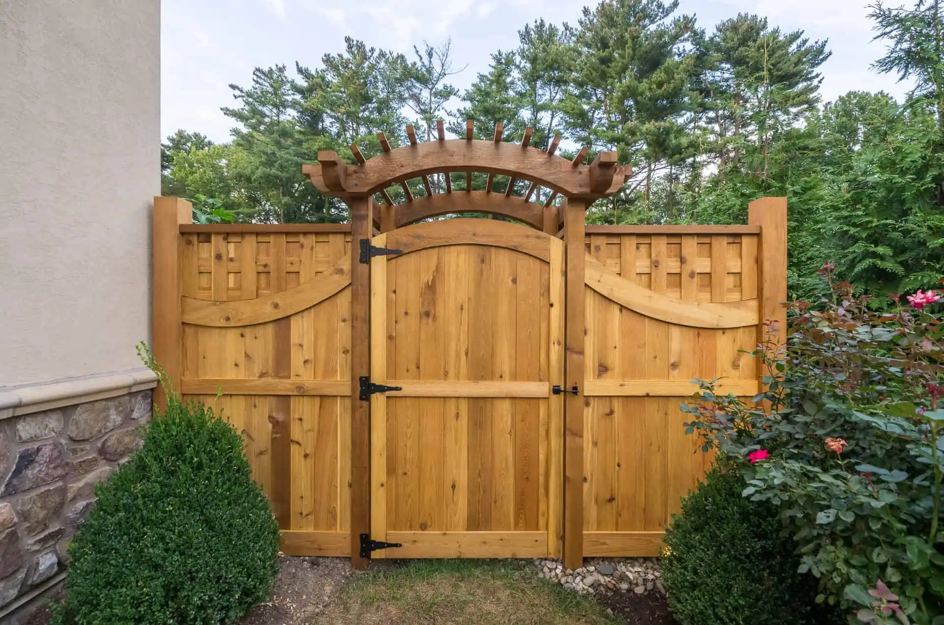 Wooden garden gate with an arched top and decorative trellis design, flanked by wooden fencing. Shrubs and flowering plants grow in front, with trees and blue sky in the background.