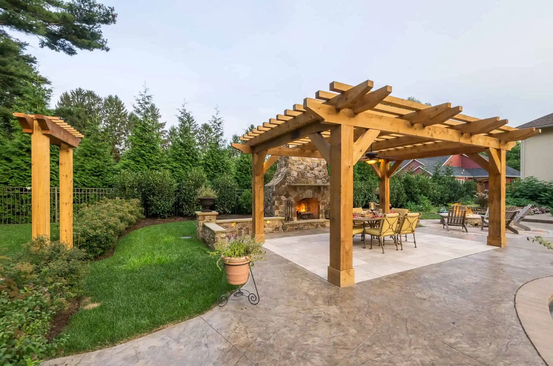 A wooden pergola with a dining area and fireplace sits on a stone patio surrounded by lush green grass, shrubs, and trees, creating an inviting outdoor living space.