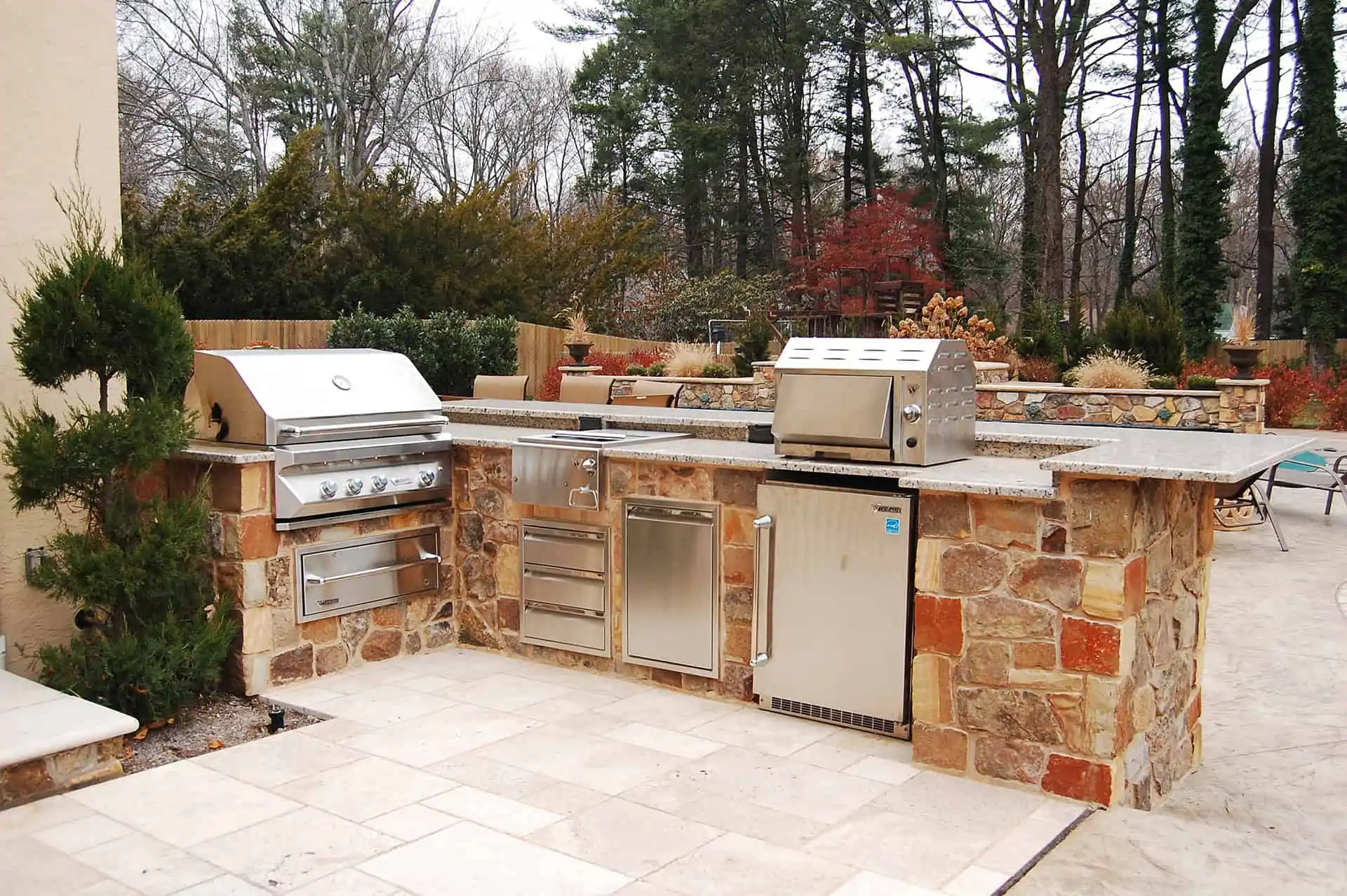 Outdoor stone kitchen with built-in stainless steel grill, smoker, storage drawers, and mini fridge, surrounded by trees and plants; light-colored tile flooring in the foreground.