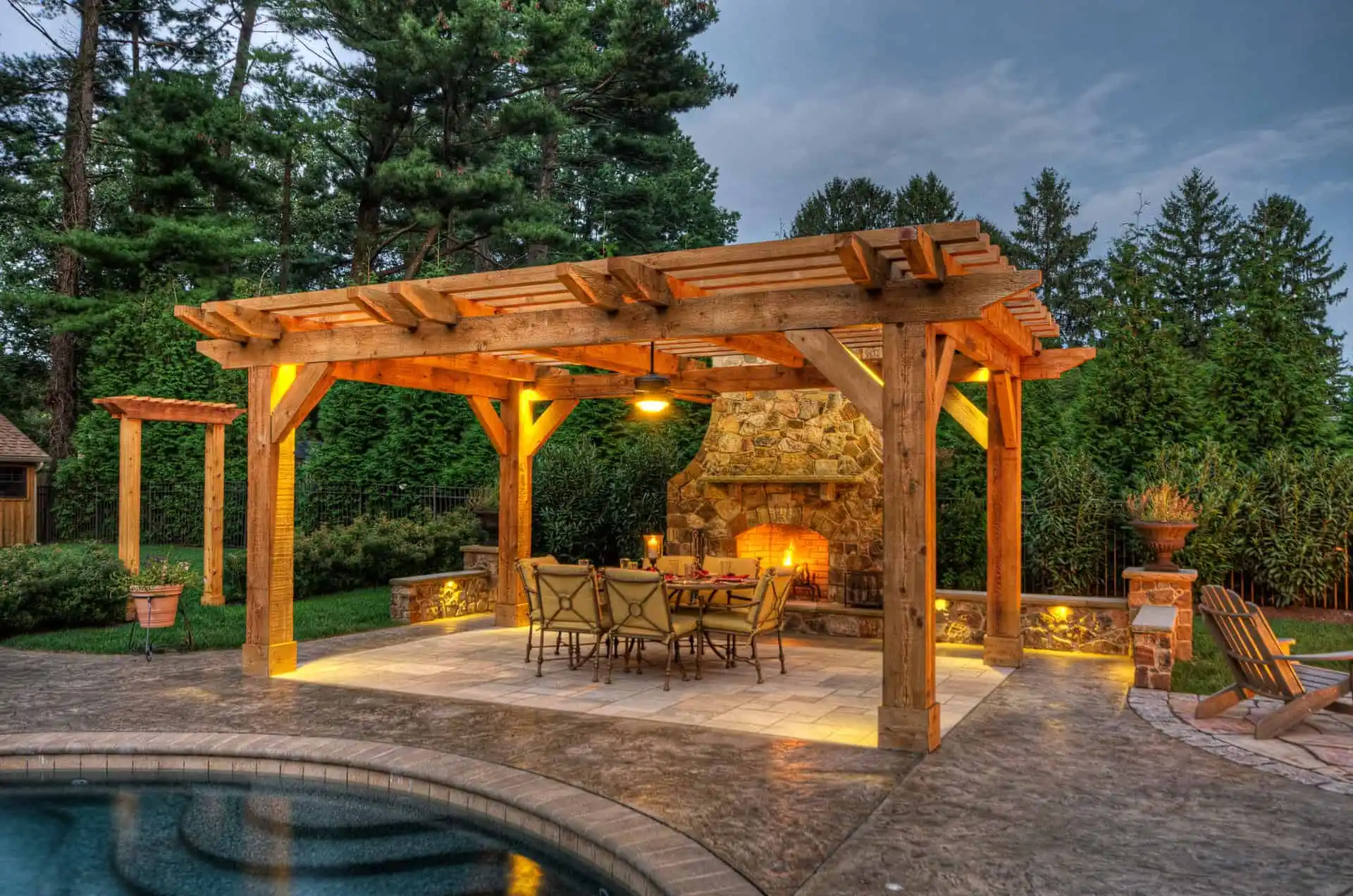 A wooden pergola with warm lighting shelters a dining table and chairs on a stone patio, beside a pool and an outdoor stone fireplace, surrounded by lush trees and greenery at dusk.