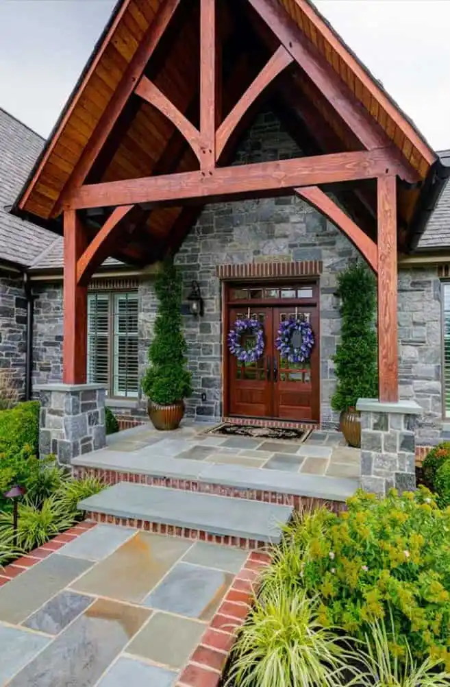 A stone house with a wooden gabled porch, double wooden doors decorated with purple wreaths, two potted plants, and landscaped greenery along a stone pathway.