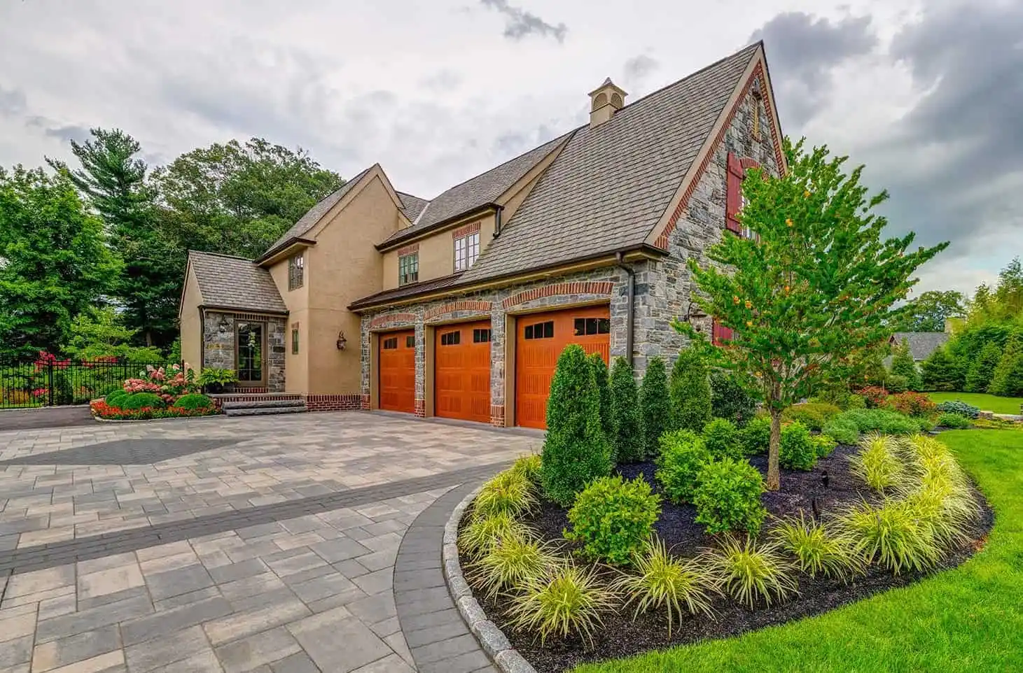 A large, elegant stone and stucco house with three wooden garage doors, manicured landscaping, vibrant green grass, and a paved driveway under a cloudy sky.