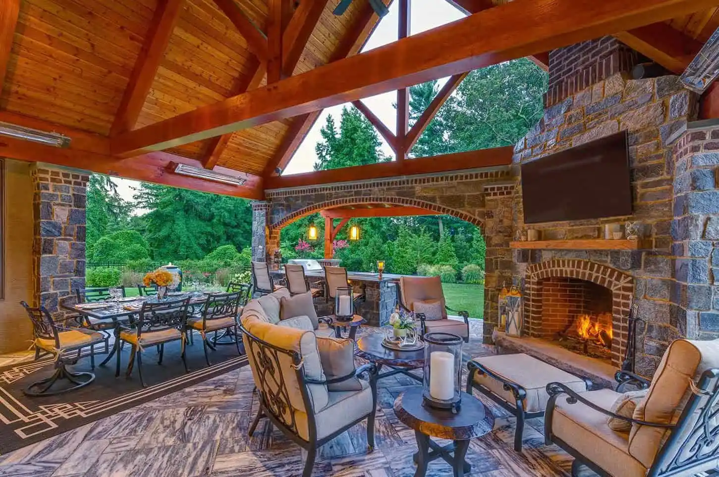 Spacious covered patio with wooden ceiling, stone fireplace, TV, cushioned seating, dining table, and outdoor lighting. Large windows open to a lush green garden with trees and shrubs. Cozy fire burns in the fireplace.