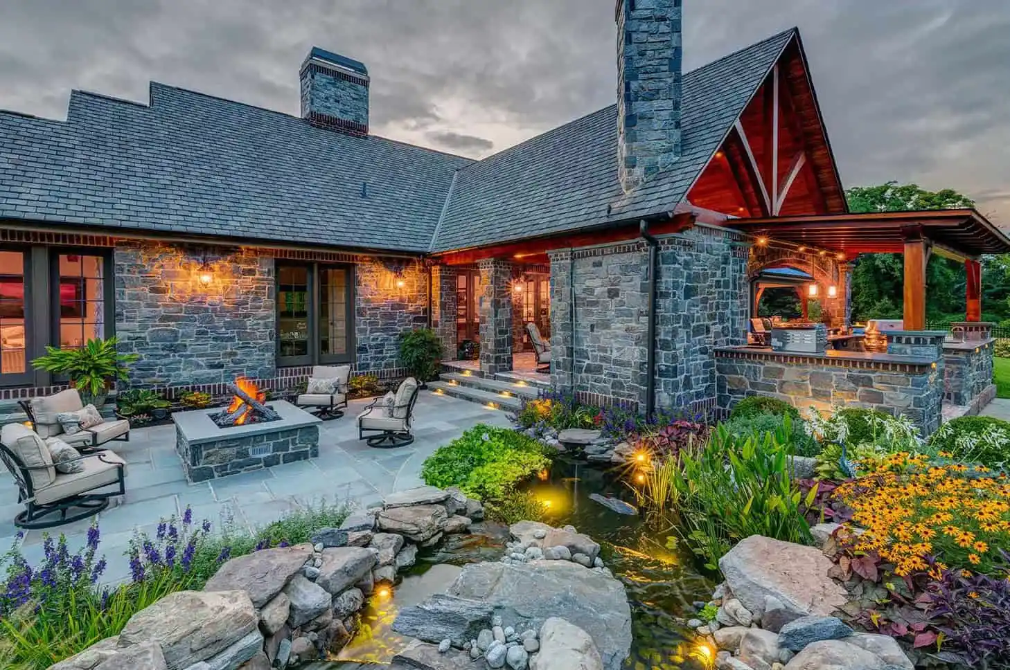 A stone house with warm outdoor lighting, a furnished patio, fire pit, and landscaped garden featuring a small pond with rocks and lush greenery at dusk.