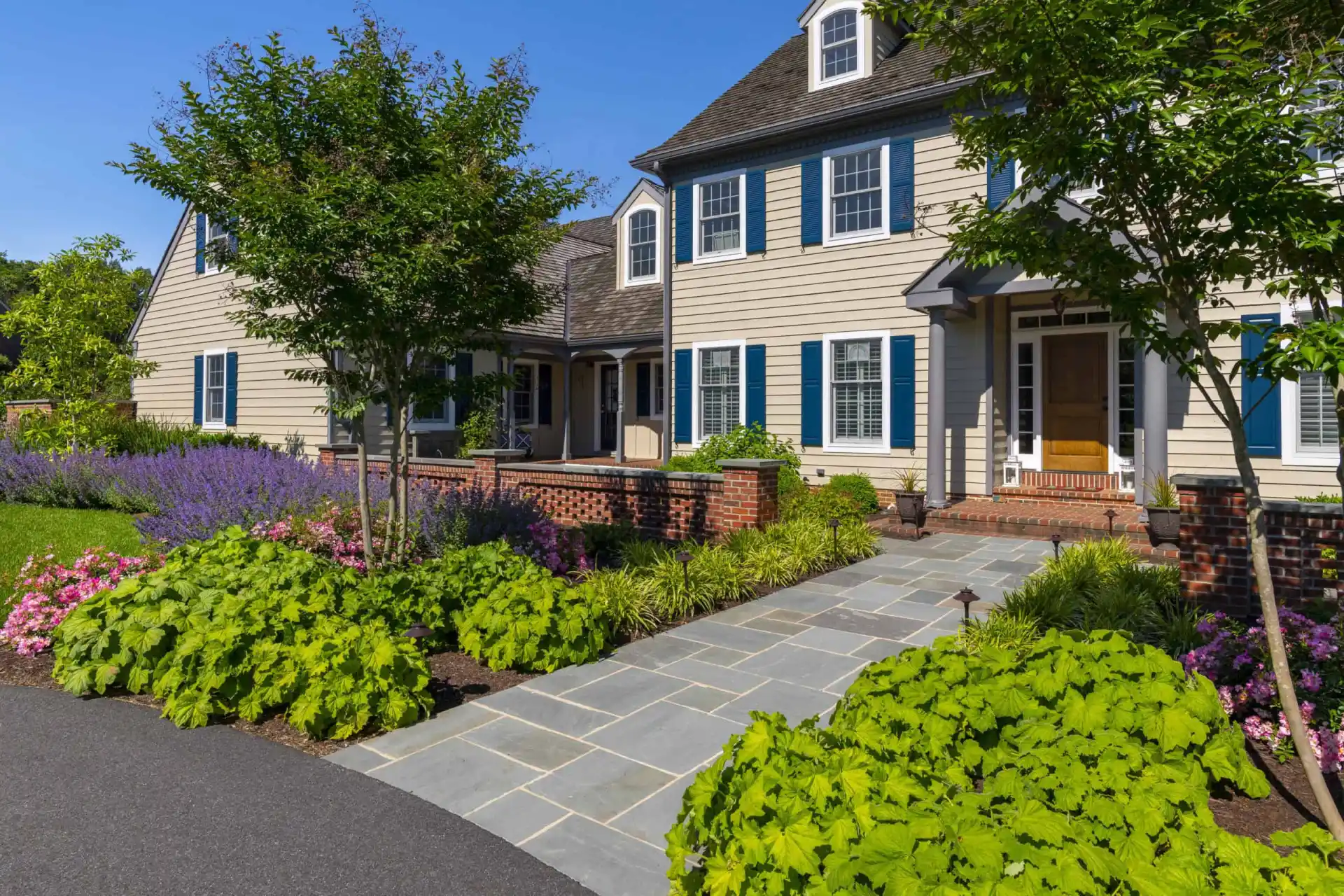 A well-maintained house with tan siding, dark blue shutters, and white trim. A stone walkway leads to the front door, bordered by lush green plants, flowers, and a brick garden wall under a clear blue sky.