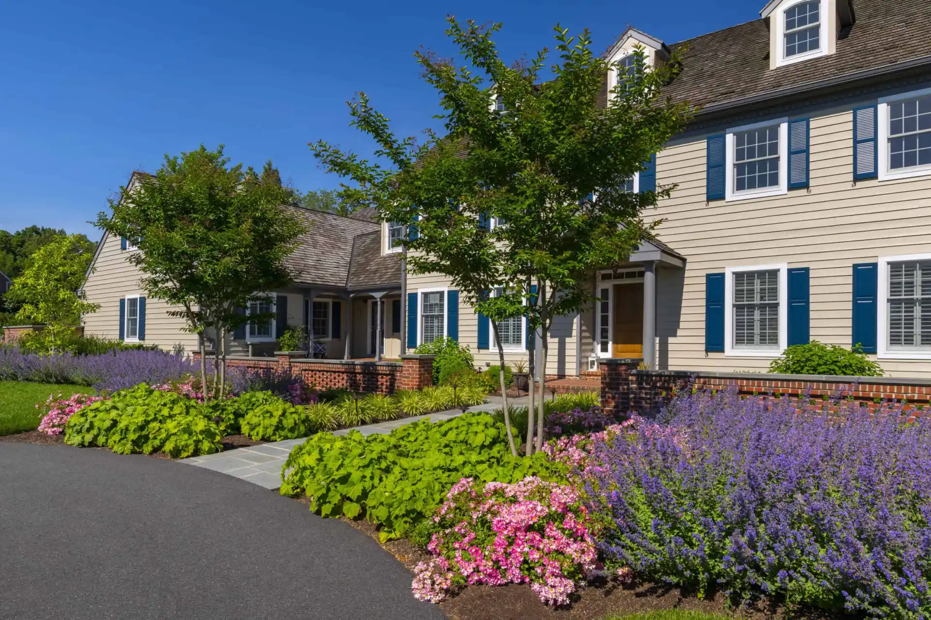 A large beige house with blue shutters is surrounded by vibrant landscaping, including green bushes, pink and purple flowers, and small trees, with a pathway leading to the entrance under a clear blue sky.