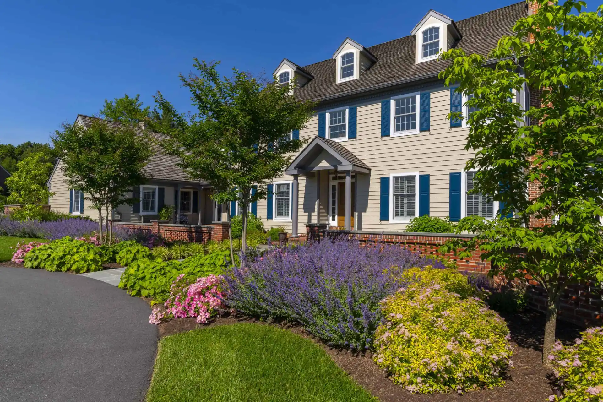 A large, two-story house with beige siding, blue shutters, and dormer windows, surrounded by lush landscaping, blooming pink and purple flowers, green bushes, and a neatly edged driveway under a clear blue sky.