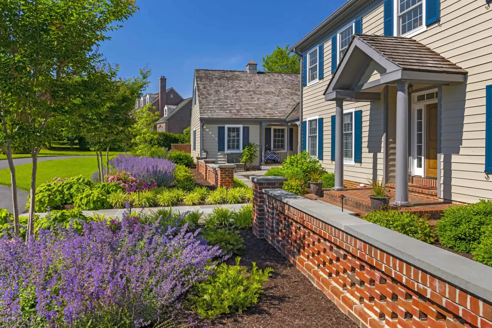 Front yard of a house with a well-maintained garden, blooming purple flowers, green shrubs, a brick walkway, and a brick wall. The house has beige siding, blue shutters, and a gray shingle roof under a sunny sky.