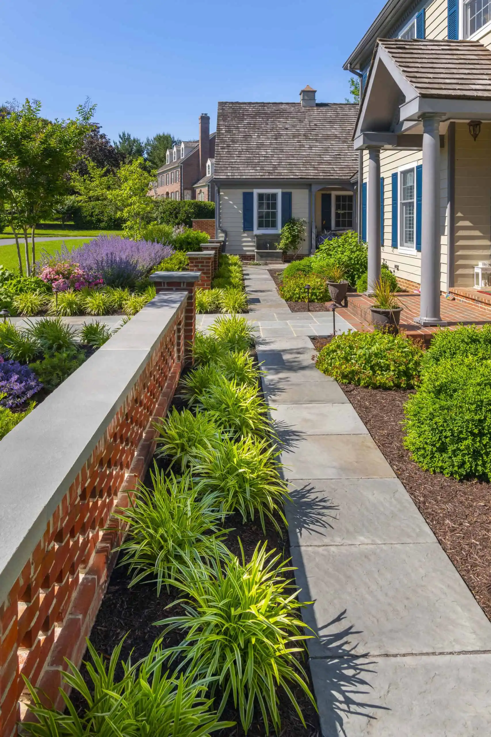 A neatly landscaped front yard with a stone walkway, low brick wall, green shrubs, ornamental grasses, purple flowering plants, and a traditional house with beige siding and a covered porch.