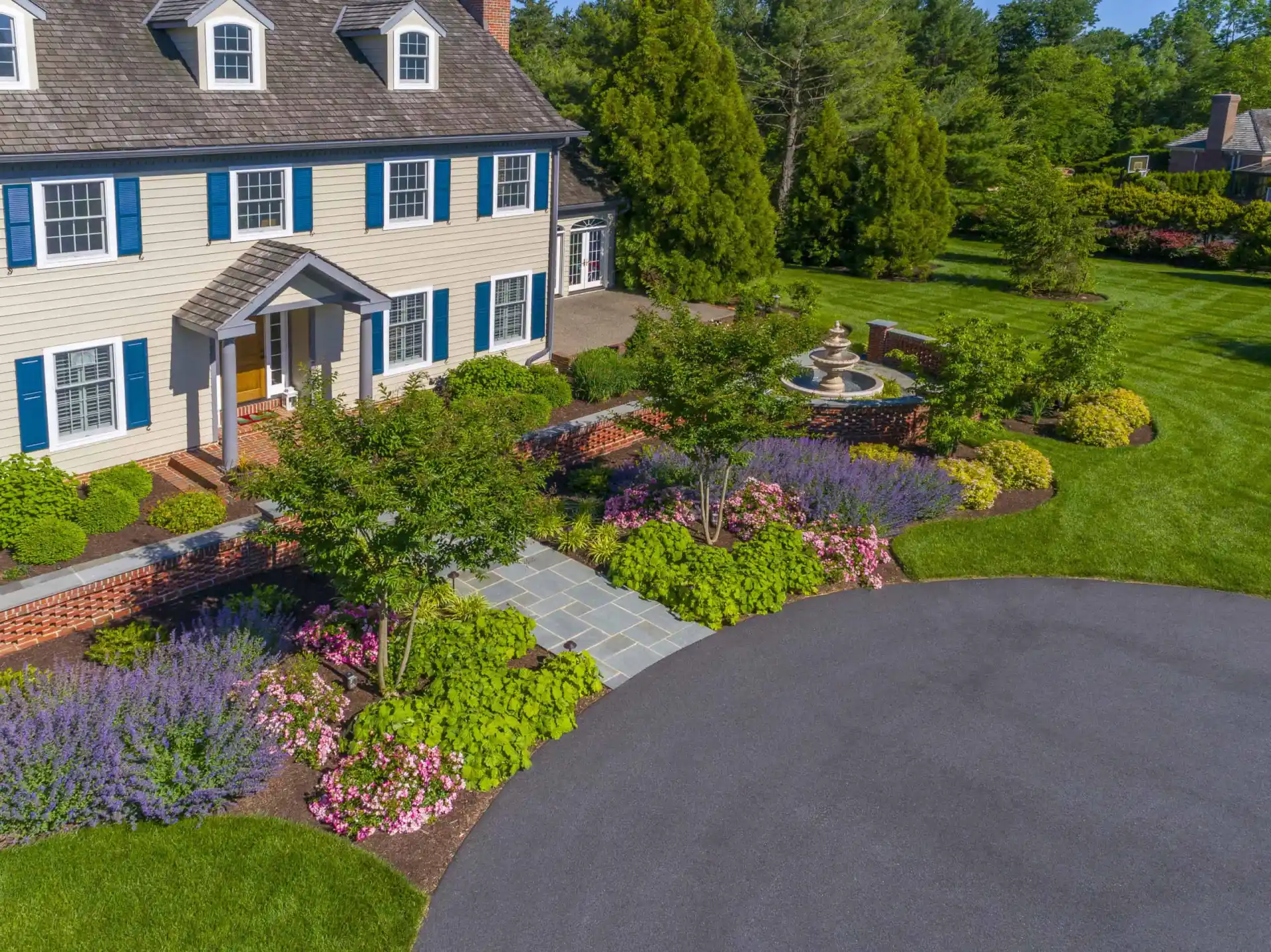 Aerial view of a large house with blue shutters, surrounded by manicured gardens, colorful flowers, green shrubs, a stone walkway, and a circular driveway with a fountain in the yard.