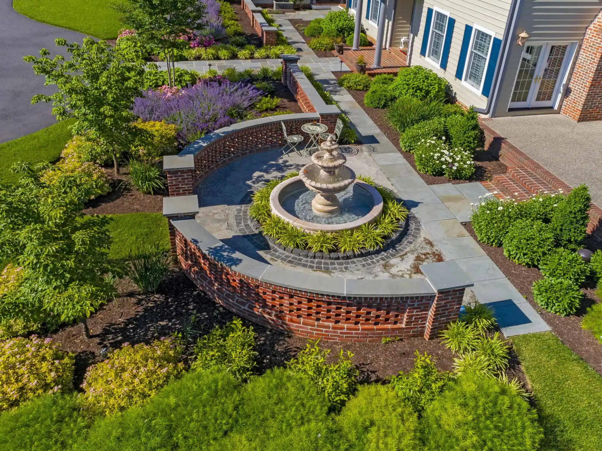 Aerial view of a landscaped garden with a circular stone fountain, curved brick wall, paved walkway, green shrubs, flowering plants, and a patio area beside a building with blue shutters.