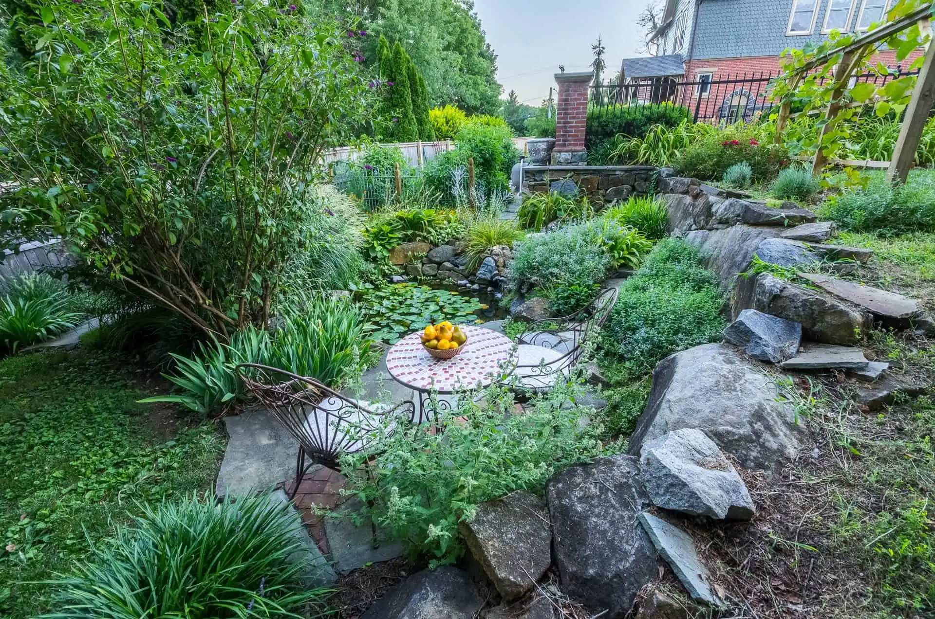 A small round table with a bowl of lemons and two chairs sits in a lush garden surrounded by greenery, rocks, and a pond with lily pads; a house and fence are visible in the background.