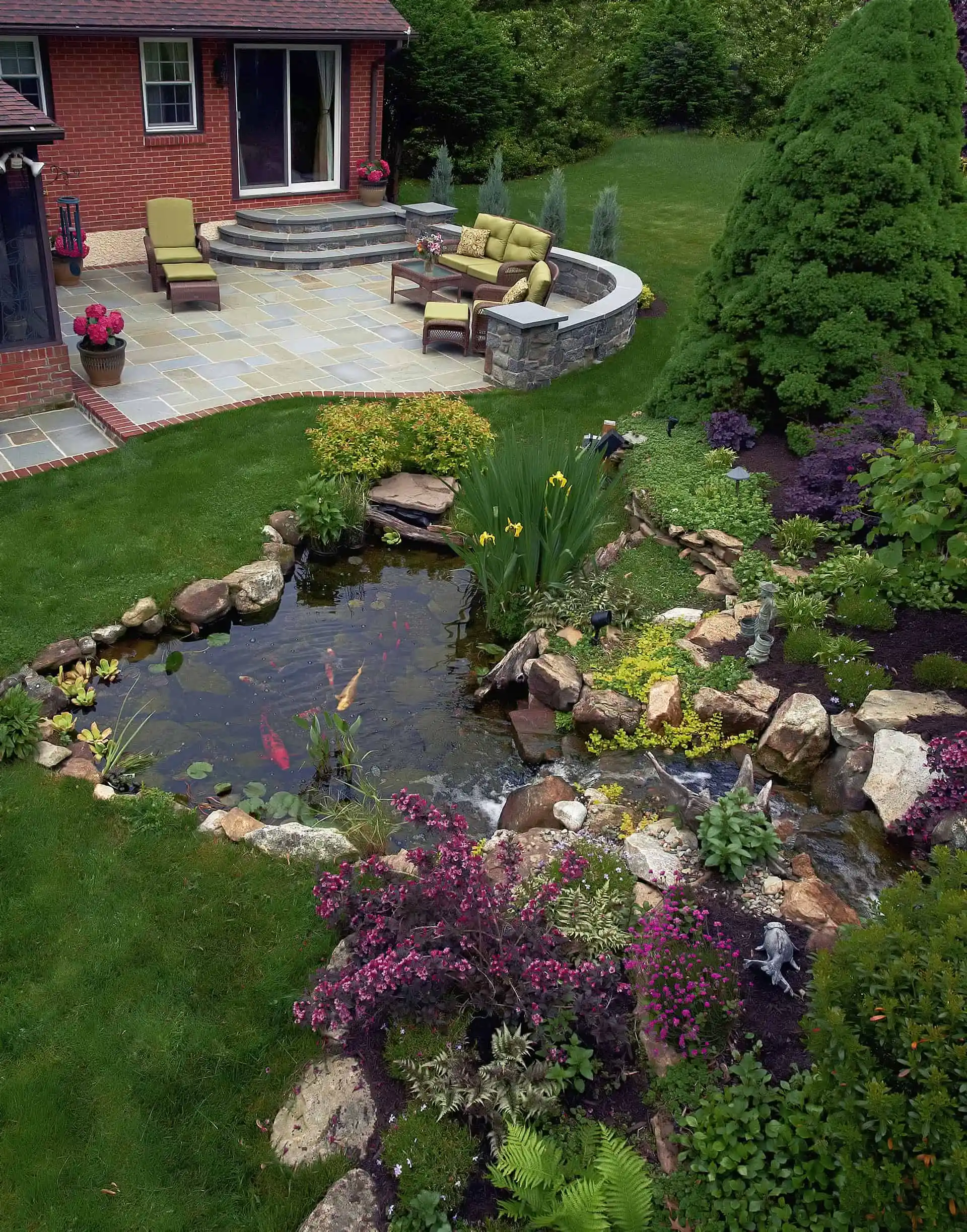 An aerial view of a landscaped backyard with a stone patio, outdoor seating, vibrant flower beds, a small pond with koi fish, and lush green grass surrounded by trees and shrubs.Water Feature and Landscaping