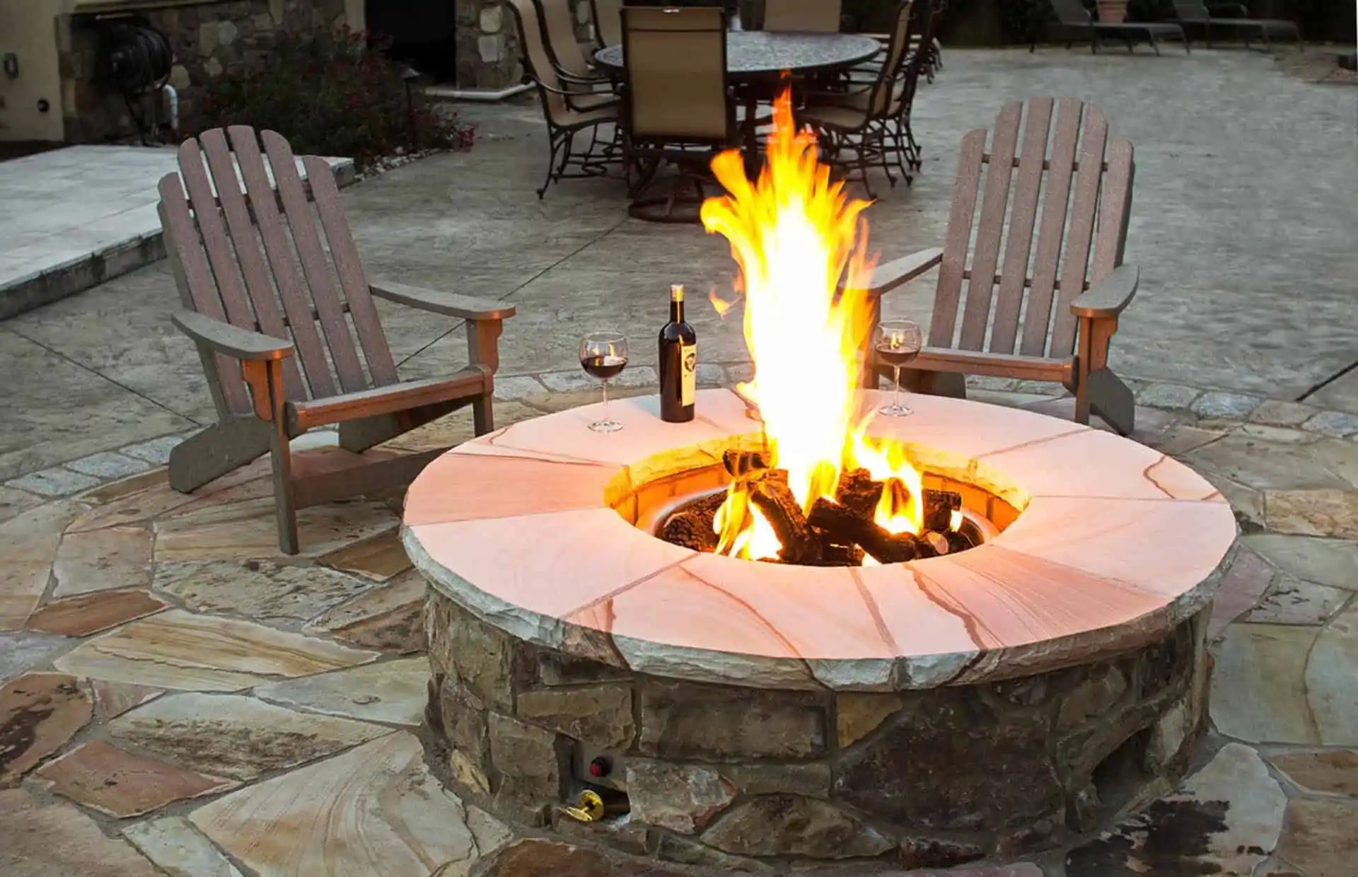 A stone fire pit with a bright flame is surrounded by two wooden chairs. There are two glasses of red wine and a wine bottle on the fire pit’s edge, set on a stone patio in an outdoor seating area.