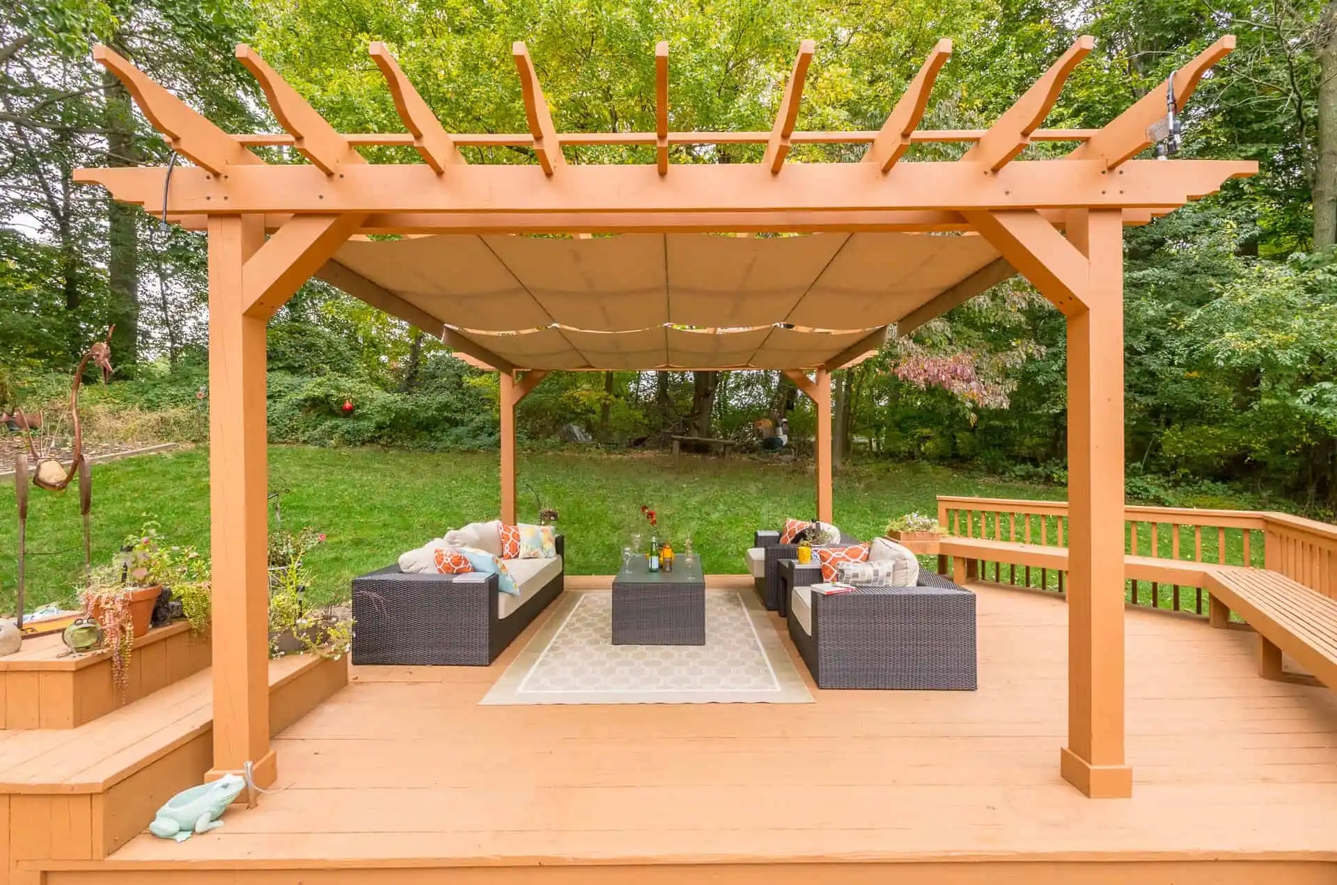 A wooden pergola with a fabric canopy covers an outdoor seating area on a deck. Wicker sofas with cushions and a central table sit on a rug, surrounded by greenery and trees in the background.