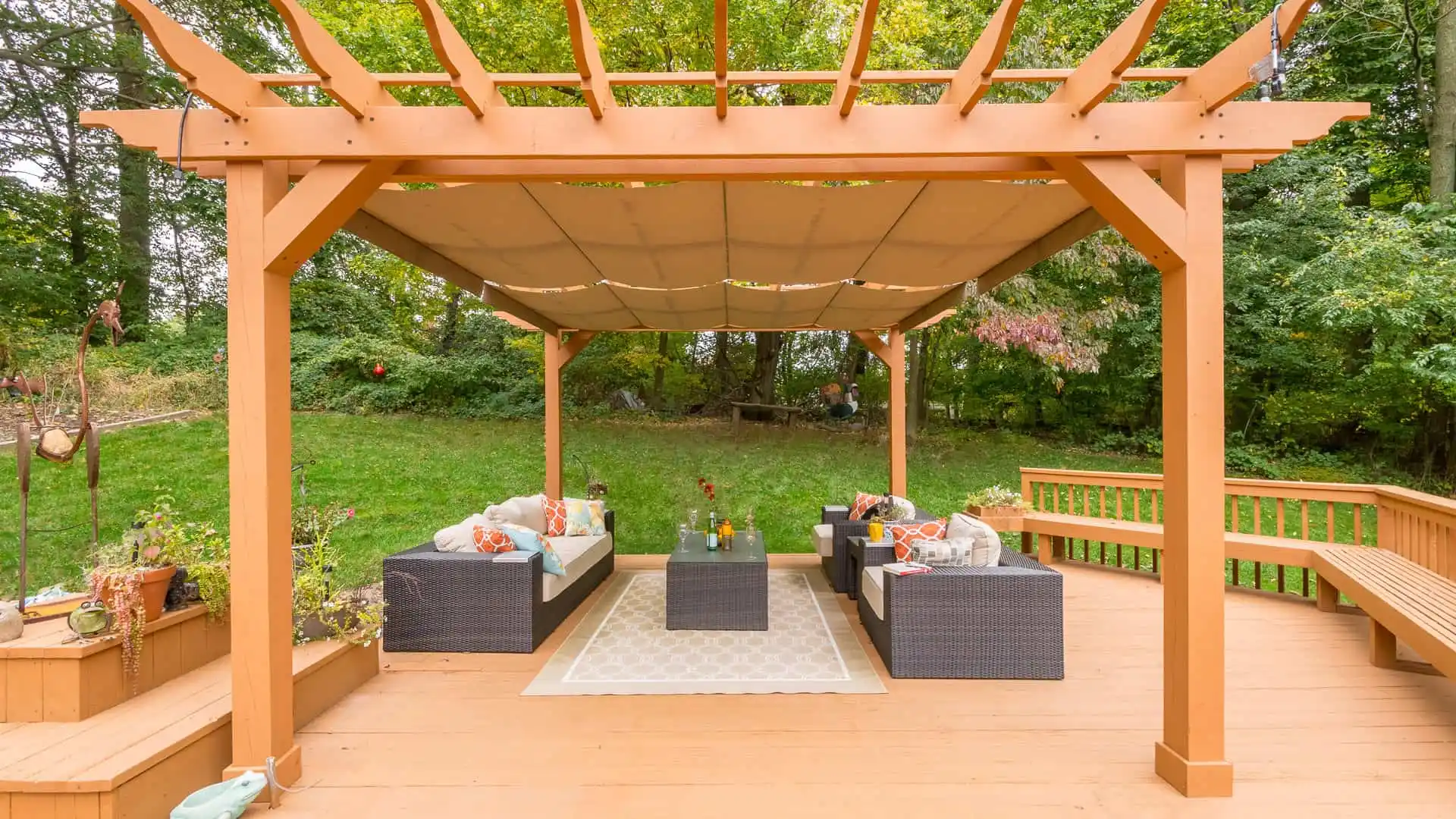 A wooden pergola with a fabric canopy shades a patio set on a deck, showcasing the essence of outdoor living in Delaware County, PA. The wicker furniture with cushions around a glass table is complemented by potted plants, all framed by lush green trees in the background.
