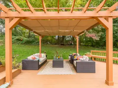 A wooden pergola with a fabric canopy shades a patio set on a deck, showcasing the essence of outdoor living in Delaware County, PA. The wicker furniture with cushions around a glass table is complemented by potted plants, all framed by lush green trees in the background.