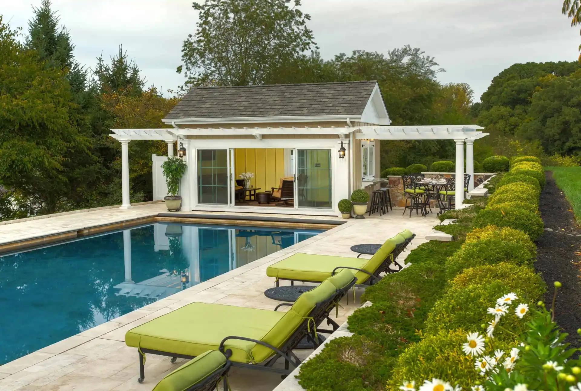 A poolside scene with green-cushioned lounge chairs, a rectangular pool, and a white pool house with sliding glass doors. There are shrubs, flowers, and outdoor seating beside the pool house.
