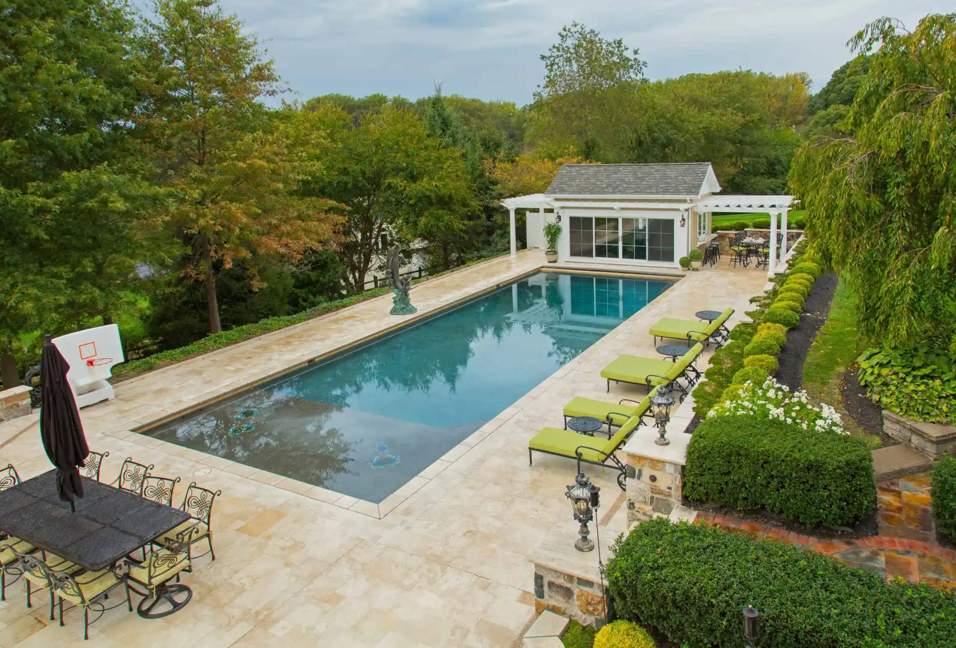 A rectangular outdoor swimming pool with lounge chairs, a pool house, a basketball hoop, and a patio dining area, surrounded by trees and landscaped greenery.