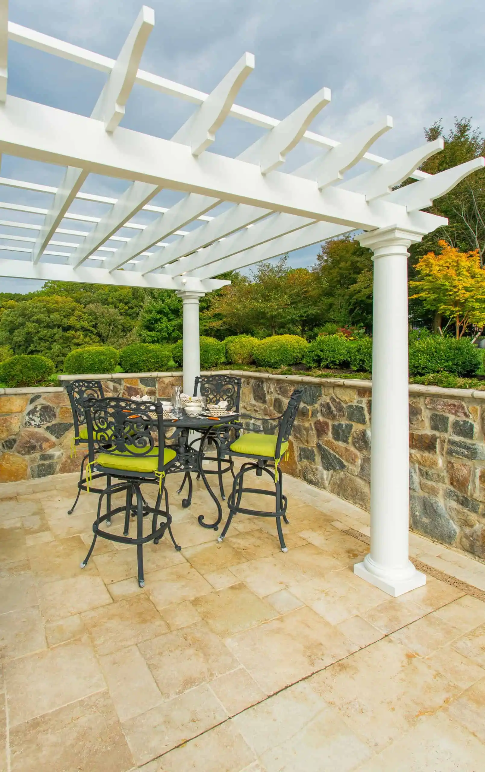 A white pergola shades a stone patio with a tall, black metal table and four chairs with green cushions. The patio is enclosed by a stone wall and surrounded by lush greenery and trees.